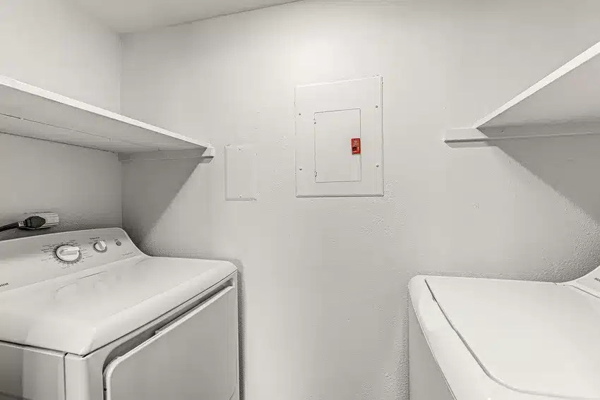 Laundry room with a white washing machine and dryer, two wall-mounted shelves, and an electrical panel on the back wall.