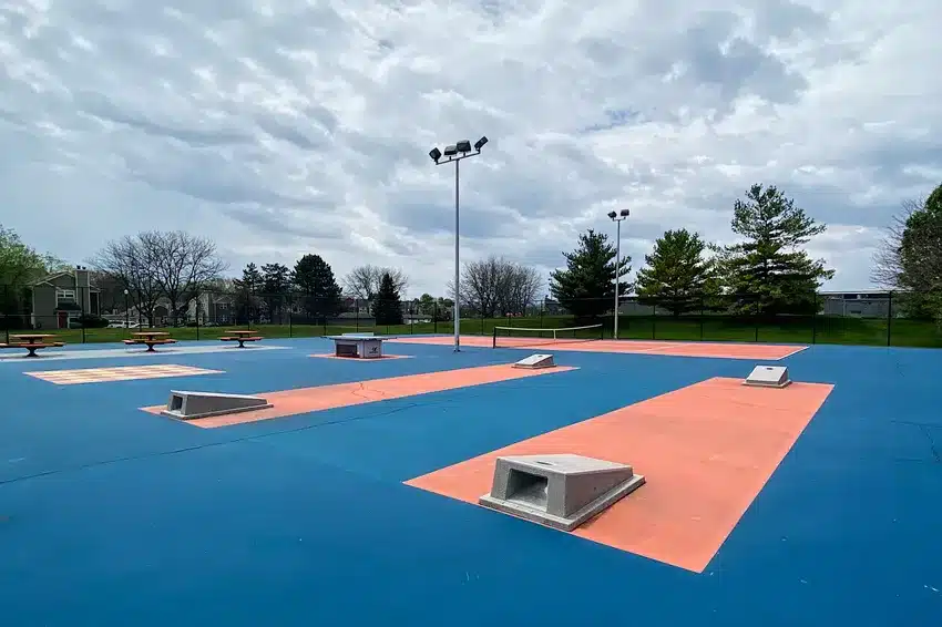 Outdoor sports court with blue and orange painted surfaces, concrete obstacles, picnic tables, and overhead lights, surrounded by trees and a fence under a cloudy sky.