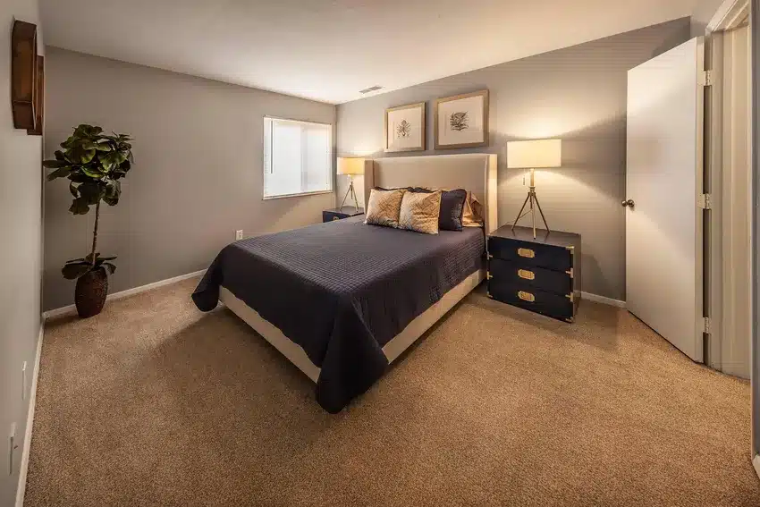 A neatly made bed with dark bedding is centered in a carpeted bedroom with two nightstands, lamps, wall art, a potted plant, and a window letting in natural light.