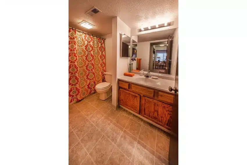 Bathroom with wood cabinets, large mirror, countertop with sink, toilet, patterned shower curtain, and tiled floor.