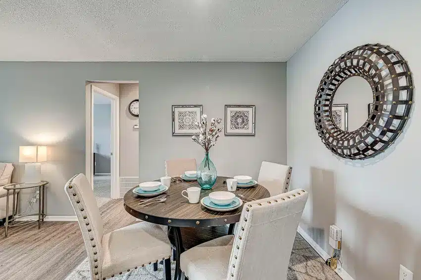 A round dining table set for four with beige chairs, a blue vase centerpiece, decorative wall mirror, and framed art in a modern dining room.