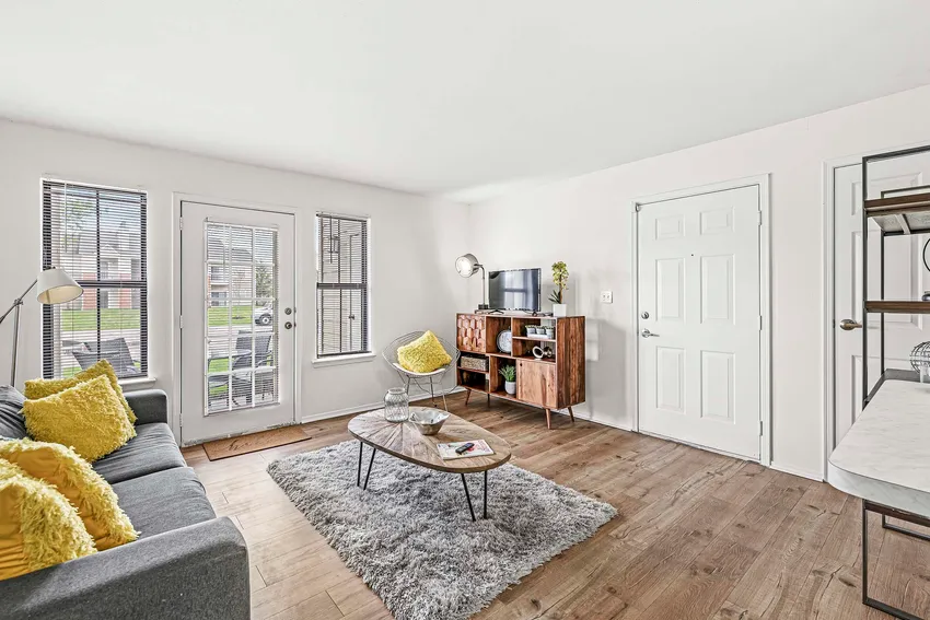 Bright living room with a gray sofa, yellow pillows, glass coffee table, TV on a wooden stand, shag rug, and wood floors. Large windows and a door let in natural light.