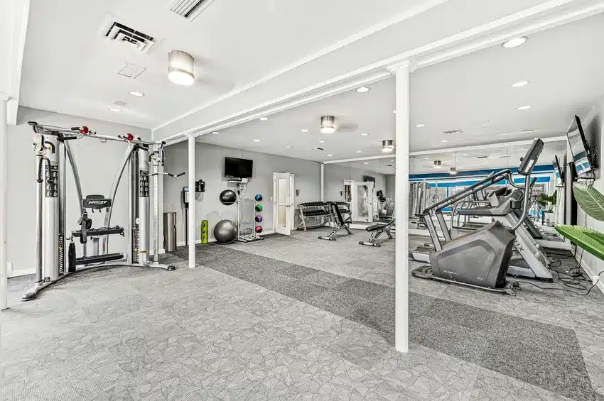 Modern gym with various fitness equipment including treadmills, a cable machine, free weights, exercise balls, and wall-mounted TVs in a bright, spacious room.