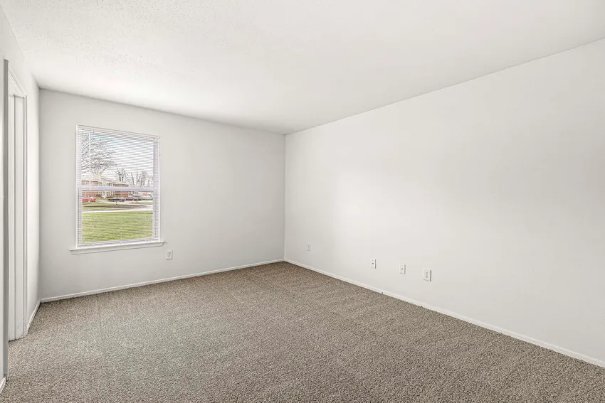 Empty room with white walls, beige carpet, a window with blinds, and electrical outlets on one wall; view of grass and trees outside.