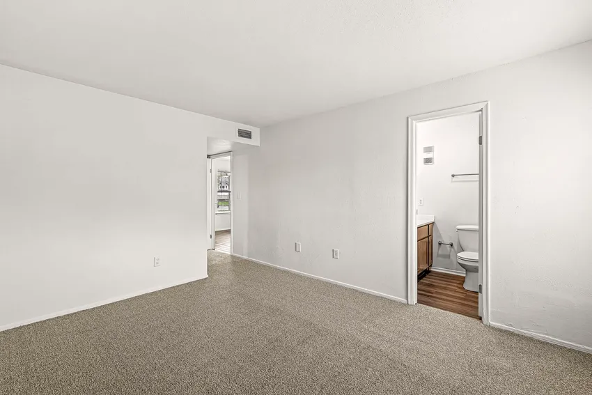 Empty room with light-colored carpet, white walls, and an open doorway leading to a small bathroom with wood flooring and visible toilet.