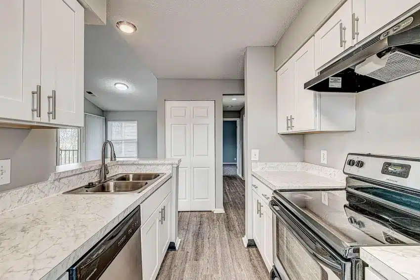 Modern kitchen with white cabinets, marble countertops, stainless steel appliances, and wood-style flooring, leading to a hallway and door with natural light from windows.