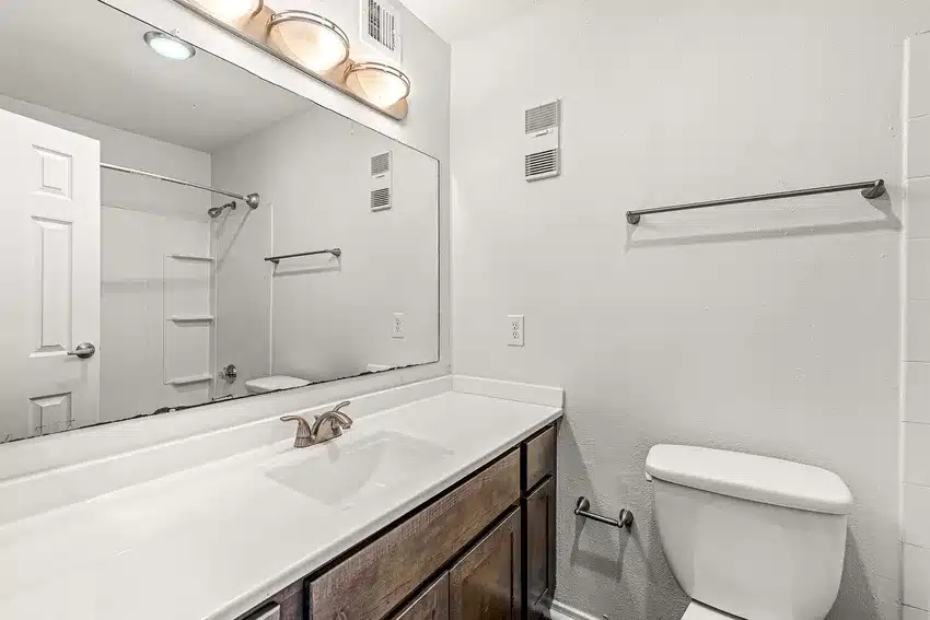 Bathroom with a large mirror above a white countertop and sink, wooden vanity, wall-mounted light fixture, and a toilet next to an empty towel bar.