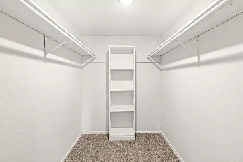 Empty walk-in closet with white walls, two hanging rods on each side, a built-in shelf unit in the center, and carpeted flooring.