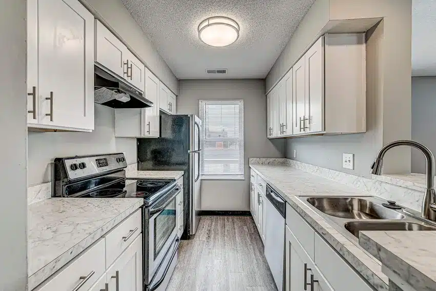 Modern galley kitchen with white cabinets, stainless steel appliances, marble countertops, and a window at the end letting in natural light.