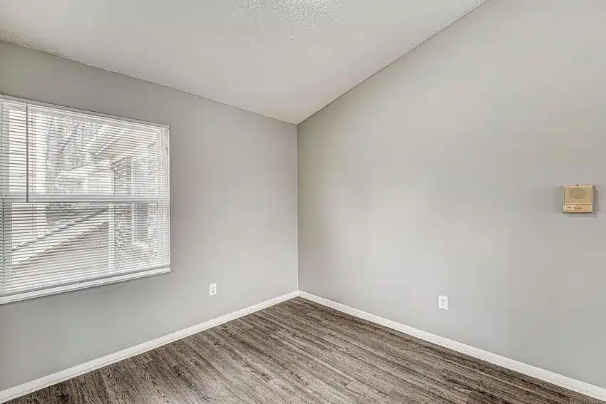 Empty room with light gray walls, a textured white ceiling, wood-look flooring, an electrical outlet, and a window with closed blinds letting in natural light.