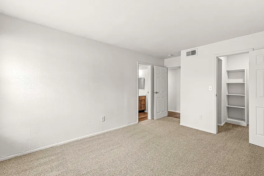 Empty carpeted room with white walls, two open doors—one leading to a bathroom with a vanity, the other to a closet with shelves. Ceiling vent and electrical outlet visible.
