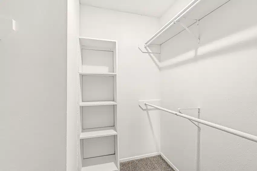 Empty walk-in closet with white walls, built-in shelves on the left, and hanging rods on two sides above a carpeted floor.