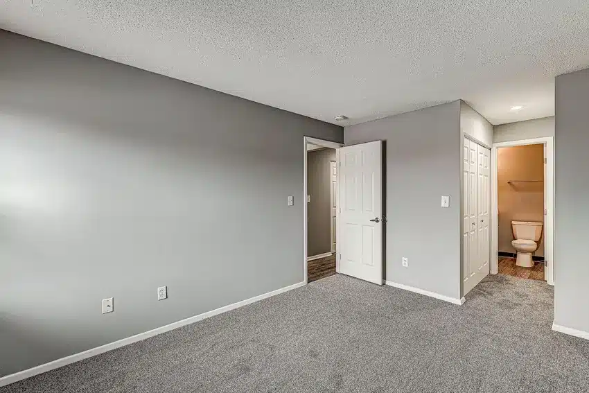 Empty carpeted room with gray walls, two open doors—one leading to a hallway, the other revealing a bathroom with a visible toilet.