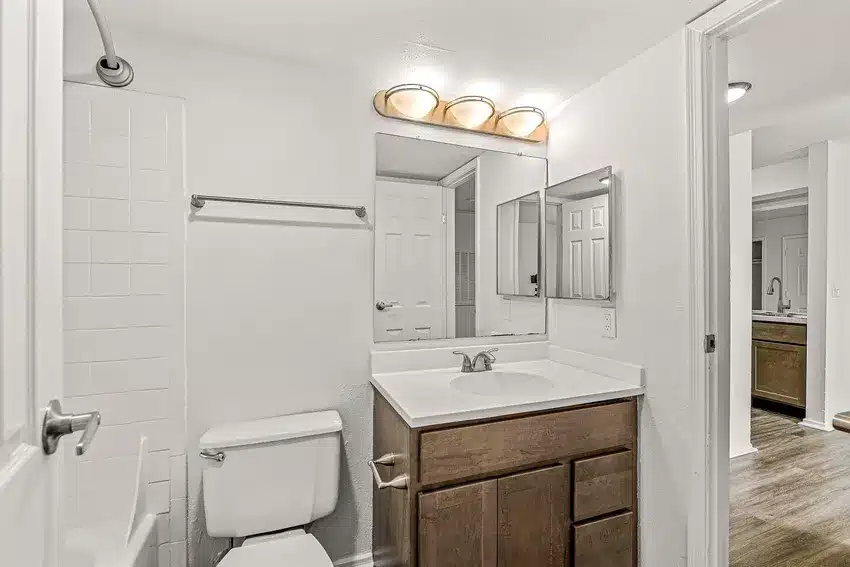 A bathroom with a wooden vanity, white countertop, rectangular mirror, toilet, towel rack, and a shower with white tile. Three-light fixture above the mirror and an open door to the hallway.