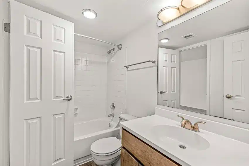 A clean, white bathroom with a bathtub-shower combo, toilet, wood vanity with sink, large mirror, and overhead lighting.