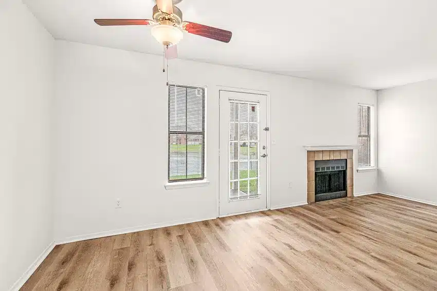 A bright, empty living room with light wood flooring, white walls, a ceiling fan, a fireplace, and glass door leading outside.
