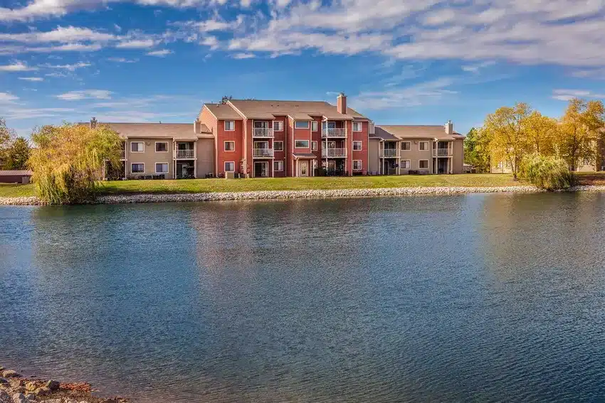 Three-story apartment building with balconies, set behind a grassy area and a pond, under a partly cloudy sky with trees on both sides.