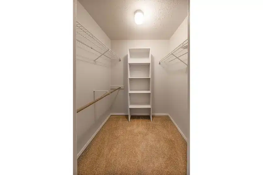 Walk-in closet with beige carpet, white walls, wire shelving on both sides, a wooden hanging rod, and a white shelving unit in the center under a ceiling light.