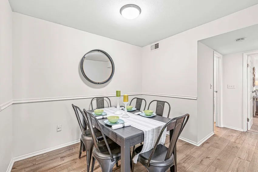 A dining room with a dark wooden table set for six, metal chairs, a striped runner, and a round mirror on the white wall. Light wood flooring and bright overhead lighting.