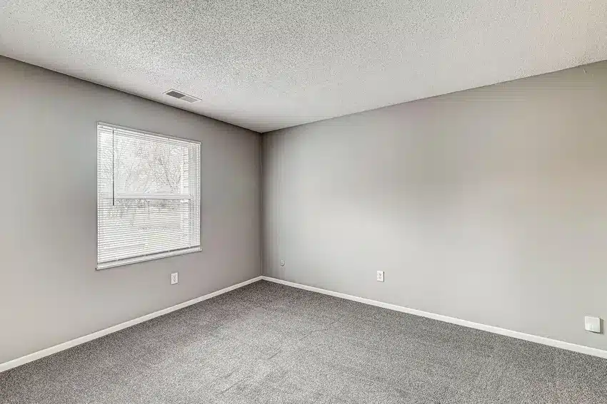 Empty room with light gray walls, a textured ceiling, a single window with blinds, and gray carpet flooring.