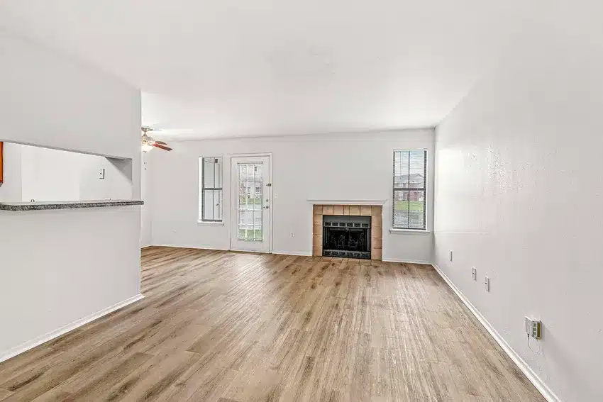 Unfurnished living room with wood flooring, white walls, a fireplace, two windows, and an open doorway to a kitchen area with a granite countertop.