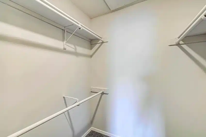 Empty walk-in closet with white walls, two levels of white wire shelves and hanging rods, and a carpeted floor. One wall shows a patch of differently colored paint.