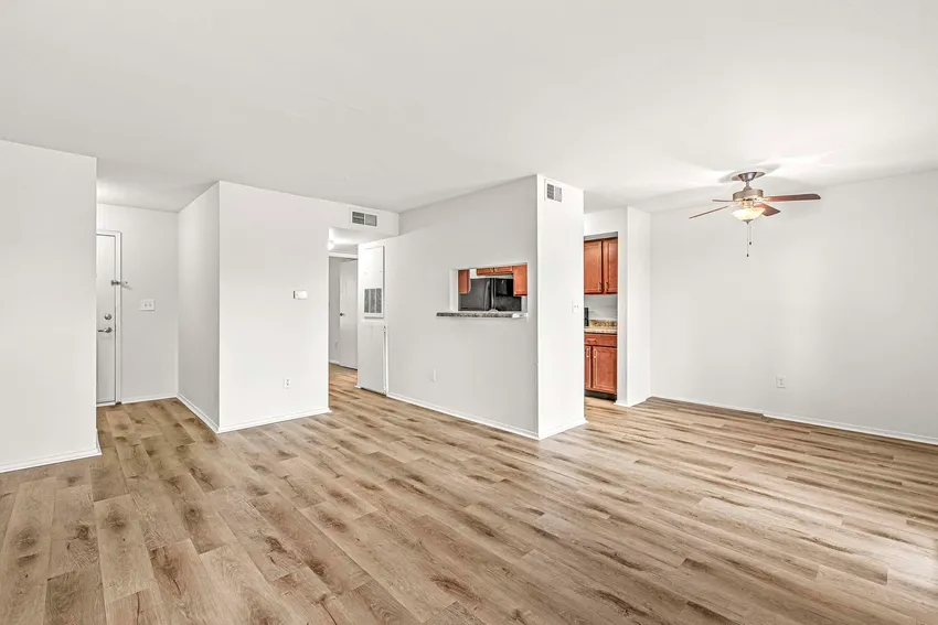 Unfurnished apartment interior with light wood flooring, white walls, a small open kitchen with dark cabinets, and a ceiling fan in the dining area.