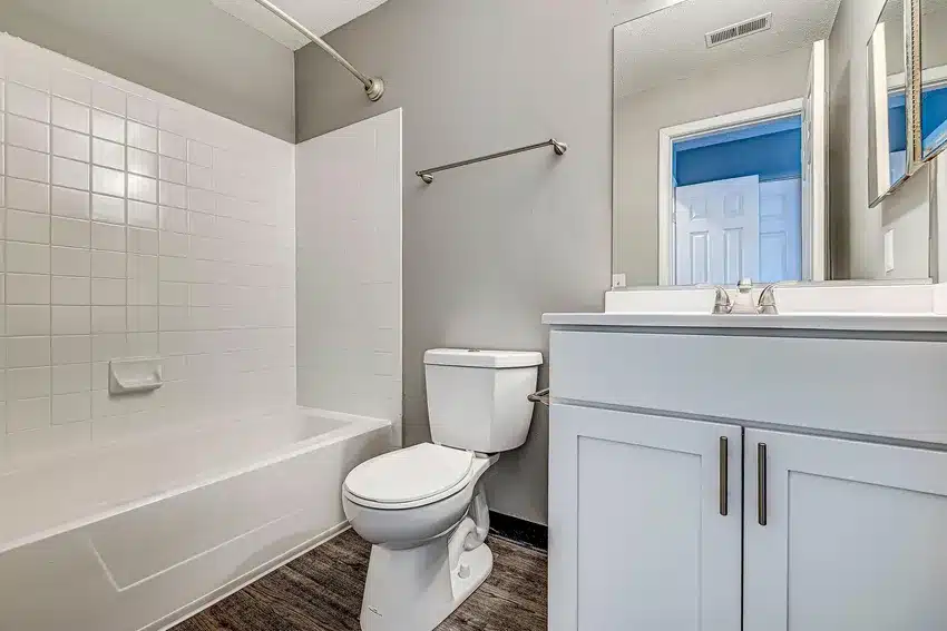 A modern bathroom with a white bathtub and tiled shower, a toilet, white vanity with sink, mirror, and towel bar on a gray wall.