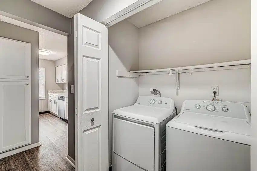 A laundry area with a white washing machine and dryer under a shelf, adjacent to a kitchen with white cabinets and wood flooring.