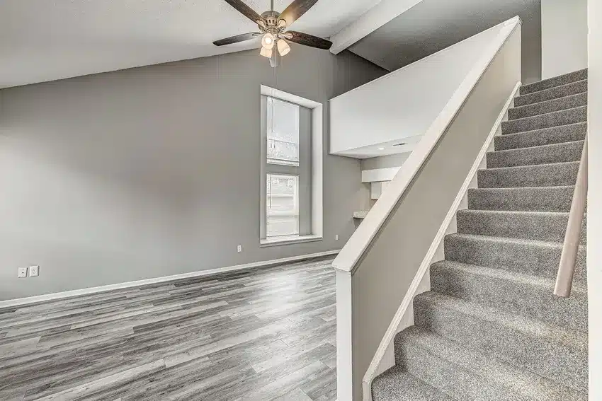 Modern living room with gray wood flooring, light gray walls, a ceiling fan, a large window, and a carpeted staircase leading to an upper level.