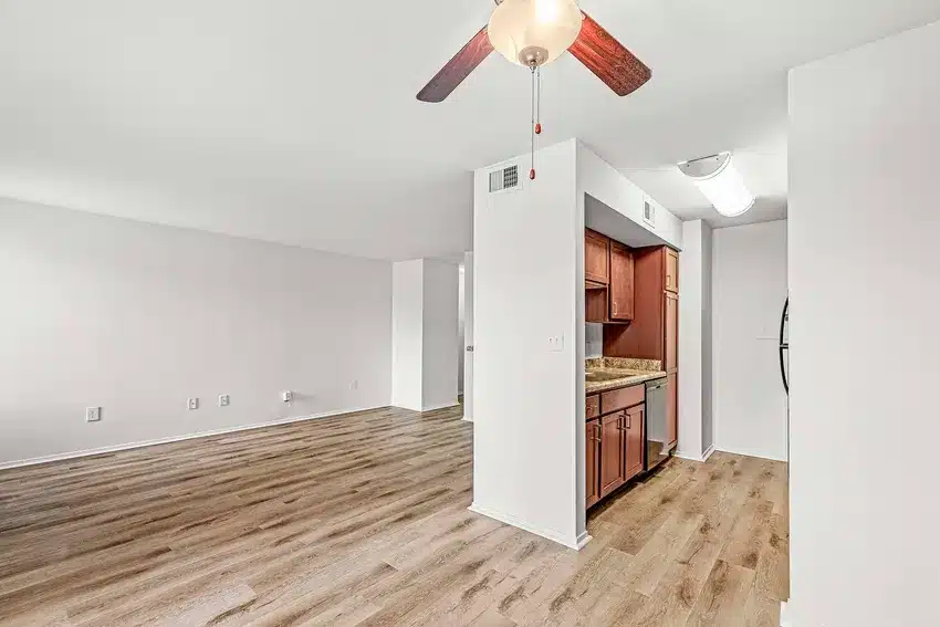 Unfurnished apartment interior with wood flooring, white walls, ceiling fan, and a small kitchen area with brown cabinets and granite counters.