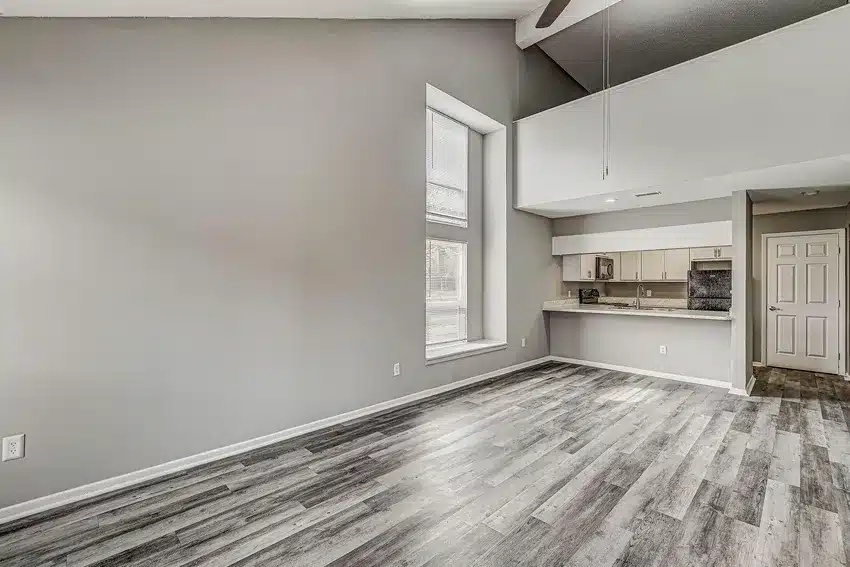Unfurnished apartment interior with gray wood-look flooring, high ceiling, large window, and open kitchen with breakfast bar.