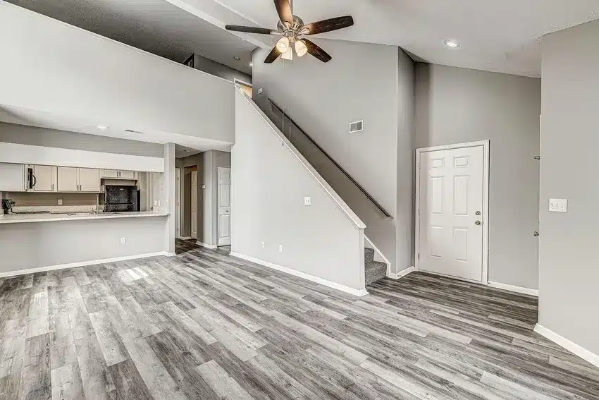 A modern, empty living area with gray wood flooring, light gray walls, a ceiling fan, stairway, and view into a kitchen with white cabinets and granite countertops.