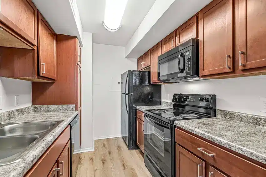 Modern galley kitchen with wood cabinets, black appliances, granite countertops, double sink, and light wood flooring under bright ceiling lighting.