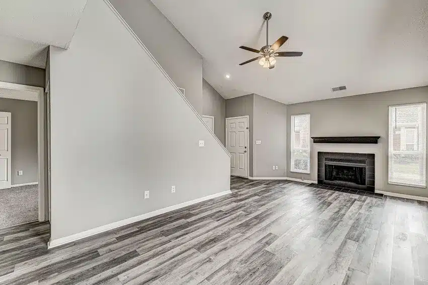 A modern living room with gray wood flooring, light gray walls, a black fireplace, tall windows, and a ceiling fan; staircase and multiple doors visible.