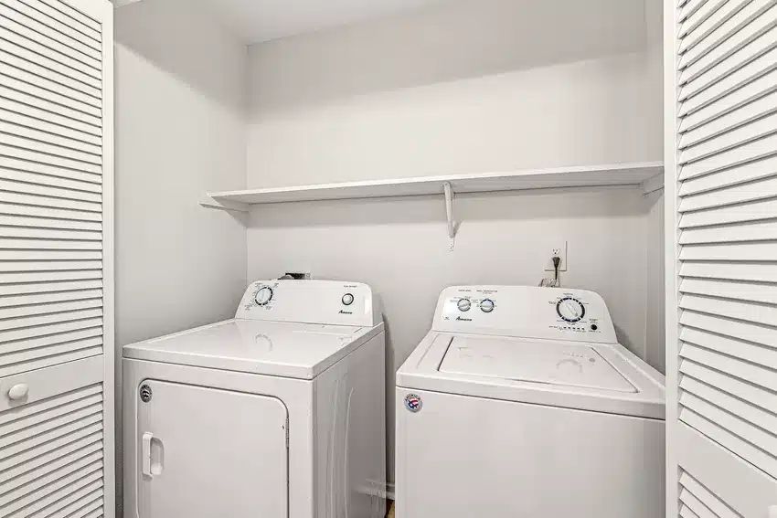 A laundry closet with a white dryer and washing machine side by side, a shelf above them, and louvered doors on either side.