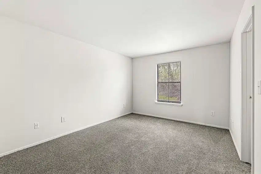 Empty room with white walls, a single window with blinds, and gray carpet flooring. The room appears well-lit with natural light from the window.