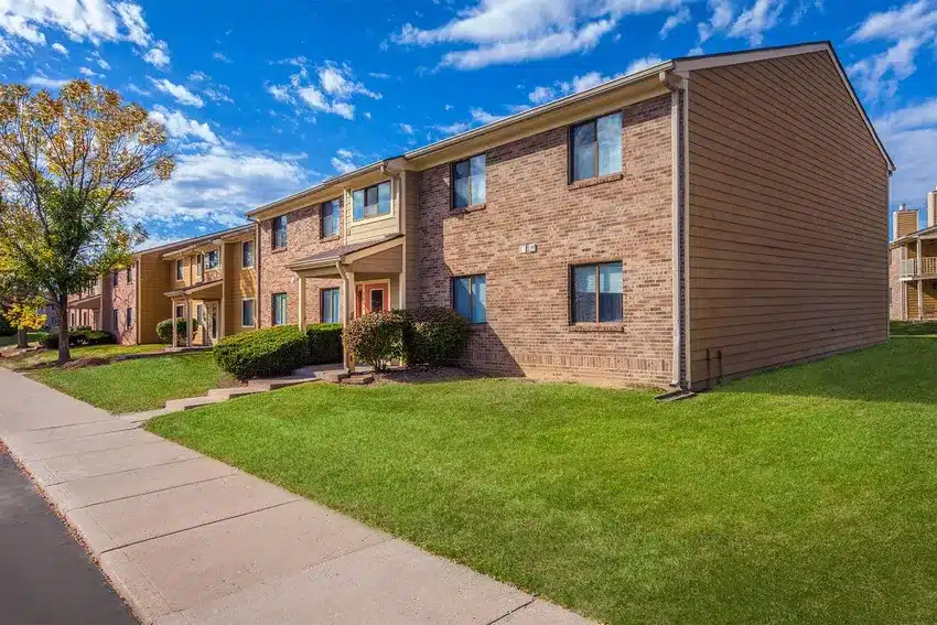 Two-story brick and siding apartment building with green lawn, shrubs, and a sidewalk under a partly cloudy sky.