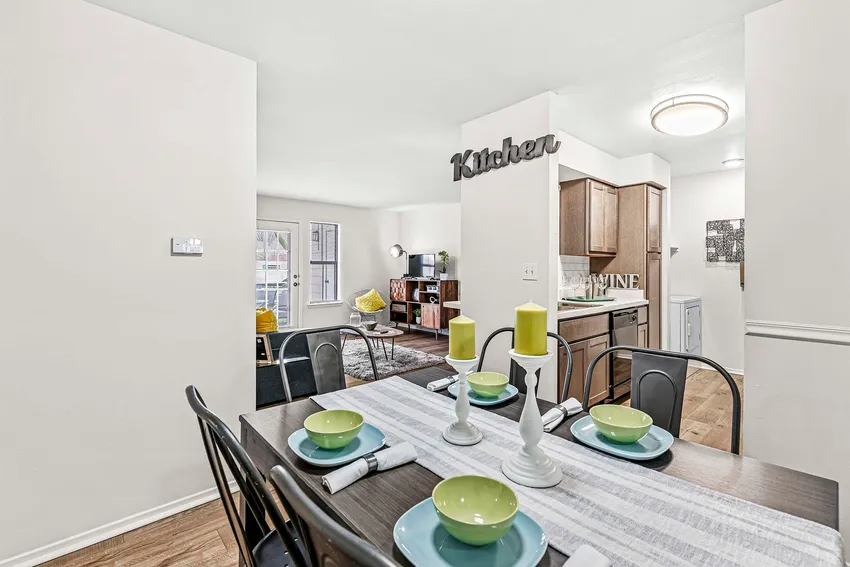 A dining area with a set table for four, green dishware, and candles, adjacent to a kitchen and an open living room with hardwood floors and modern decor.