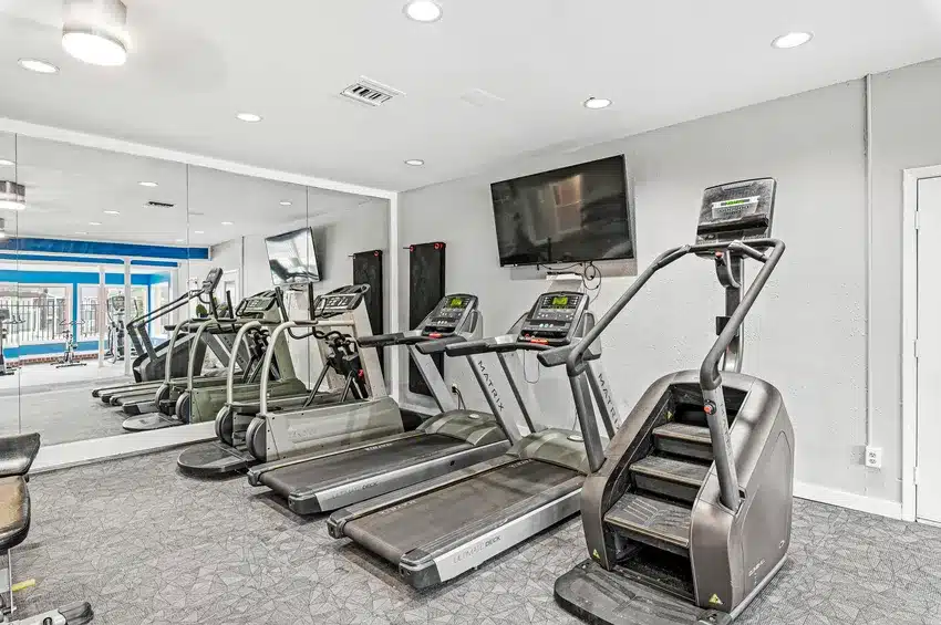 A gym room with two treadmills, a stair climber, a mirrored wall, a mounted TV, and windows showing an outdoor pool area.