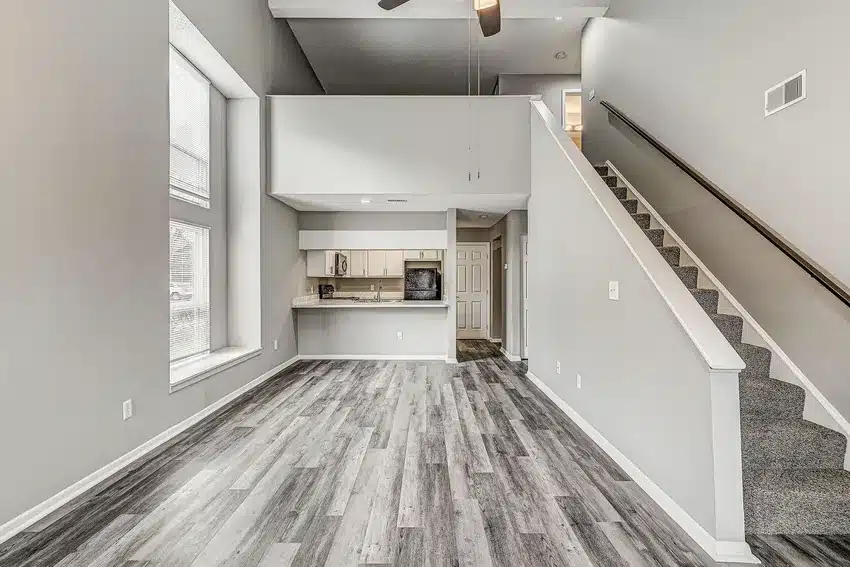 Modern loft-style living area with gray wood flooring, high ceilings, large windows, an open kitchen, and a carpeted staircase leading to an upper level.