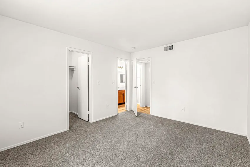 Empty, carpeted bedroom with white walls features two open doors, one leading to a closet and the other to a bathroom with a visible vanity area.