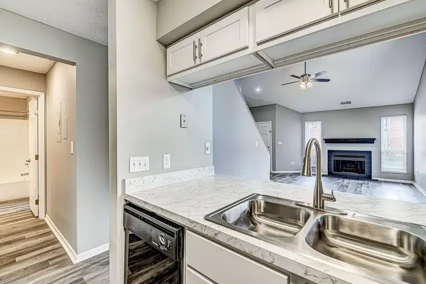 Modern kitchen with marble countertop, double sink, and stainless faucet, open to a living room with fireplace, ceiling fan, and wood-style flooring. Doorway leads to a bathroom.