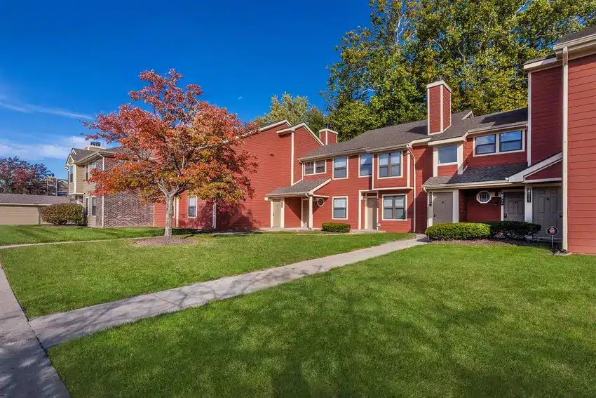 A row of red two-story townhouses with white trim, surrounded by green lawns, a sidewalk, and a tree with autumn foliage under a clear blue sky.