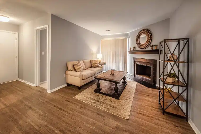 A living room with light wood floors, a beige sofa, a wooden coffee table, a fireplace with a round mirror above it, and a black metal bookshelf.
