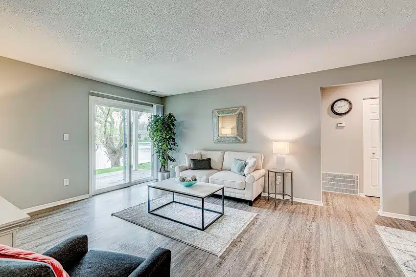 A modern living room with a white sofa, coffee table, lamp, wall art, indoor plant, wood flooring, and sliding glass doors leading outside.