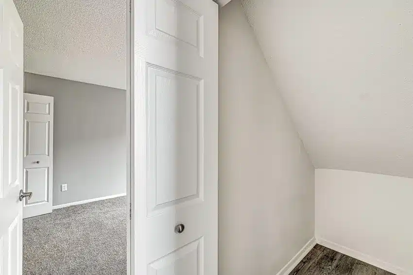 Small, empty closet space with sloped ceiling and white walls, viewed through an open door; adjacent carpeted room visible in the background.