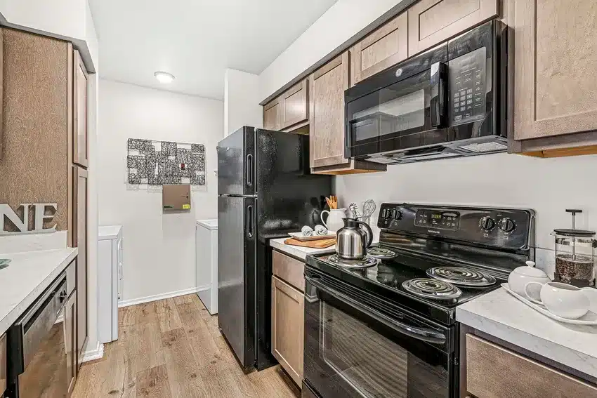 Modern kitchen with black appliances, wood cabinets, electric stove, microwave, and a stainless steel kettle; a laundry area with washer and dryer is visible in the background.