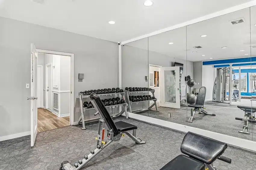 A modern gym room with dumbbells, bench presses, and a large wall mirror; open door shows hallway with wood flooring.