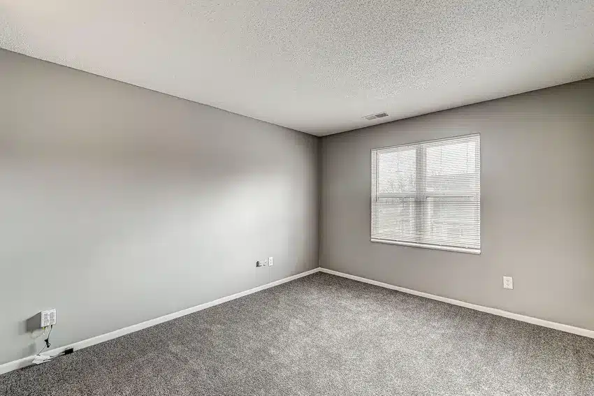 Empty room with light gray walls, a window with closed blinds, gray carpet flooring, and visible electrical outlets on the walls.